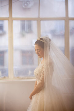 Beautiful Asian bride in white beaded wedding gown with tiara and veil, soft window light, wedding preparation portrait