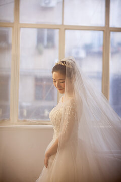 Beautiful Asian bride in white beaded wedding gown with tiara and veil, soft window light, wedding preparation portrait