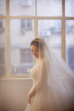 Beautiful Asian bride in white beaded wedding gown with tiara and veil, soft window light, wedding preparation portrait