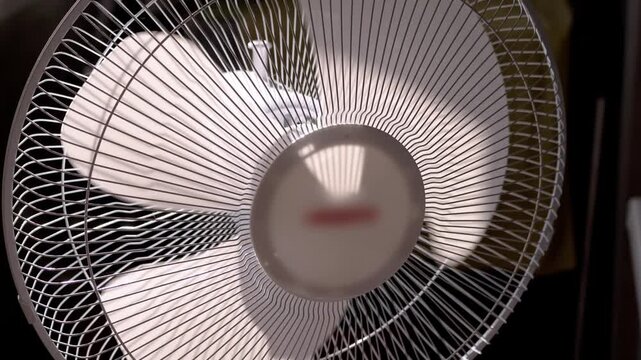 Electric fan oscillates slowly indoors during hot summer weather. Rotating plastic blades behind a wire safety grill cast rhythmic shadows to create an expressive sense of domestic relief.