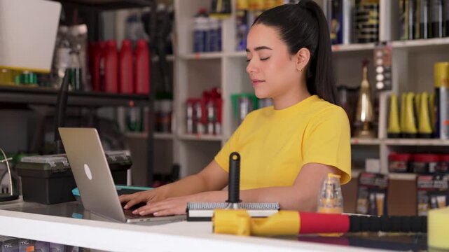 Young latina female shop assistant typing on a laptop computer behind auto parts store counter