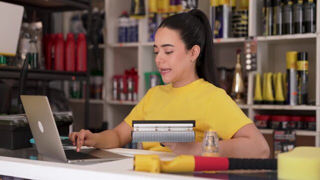 Young latina saleswoman using laptop while managing car detailing tools at store checkout counter