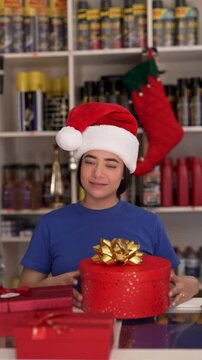 Happy latina female store clerk wearing santa hat opening red christmas gift box at checkout counter