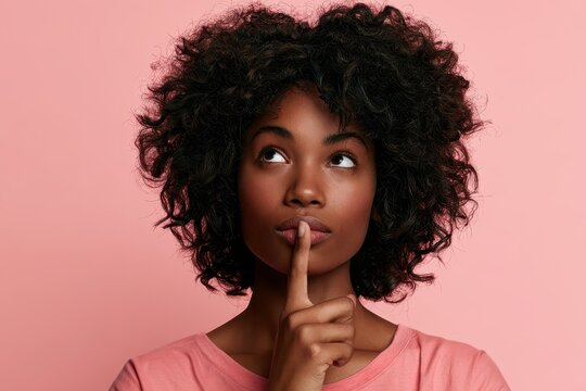 Thoughtful studio portrait: young woman in pink top with finger to lips, gazing upward against a soft pink backdrop