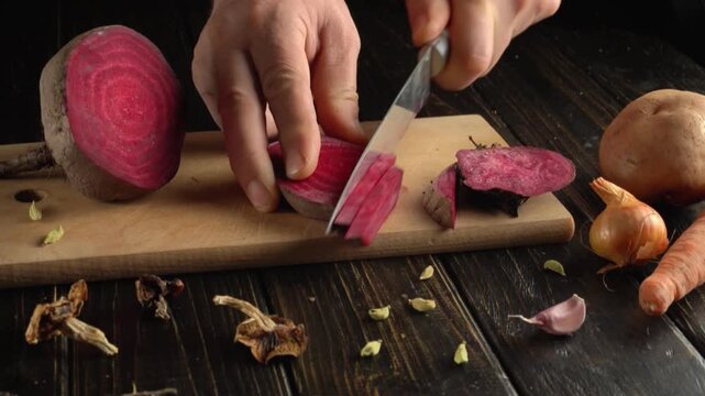 Hands slicing a red beet on a wooden cutting board with a kitchen knife, surrounded by onion, carrot, potato, garlic, and dried herbs in a rustic kitchen setting