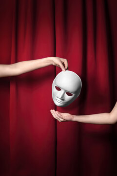 A white mask hangs between two hands against a deep red curtain, as one hand holds it above the open palm of the other. One hand drops a plain white face mask toward another waiting hand.