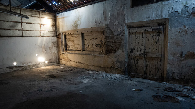 Empty abandoned warehouse interior with damaged walls, peeling paint, exposed wooden roof structure, and dusty concrete floor, showcasing building deterioration, property neglect, and urban decay.