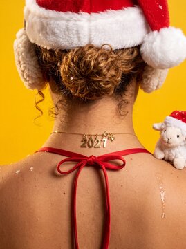 View from behind of a woman wearing a 2027 gold necklace, a red tie top, and a Santa hat, with a lamb on her shoulder