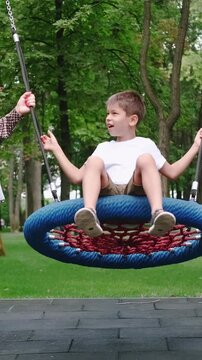 Vertical video. Family leisure. Playful childhood. Cheerful active son smiling sitting on large round swing in park talking with happy relaxed mother.