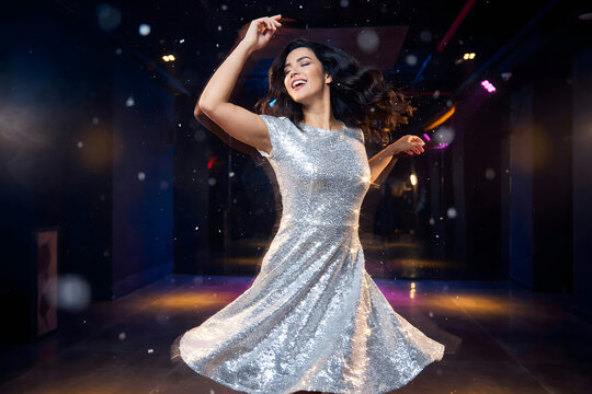 A young woman in a sparkling silver sequin dress dancing happily in a dark nightclub with colorful stage lights and floating particles in the air