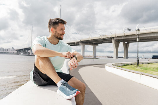 Man checks watch while relaxing by the river under a cloudy sky in a modern city