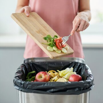 Person scraping fresh salad vegetables from a wooden cutting board into a kitchen trash bin filled with food waste