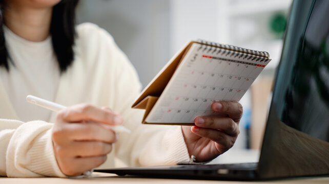 Woman using calendar to schedule meeting or appointment timetable, she holds white pen while working on laptop computer to organize daily business tasks at home office desk.