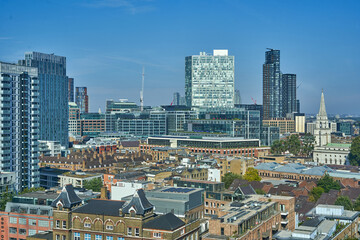 Panoramic aerial view of the London skyline showcasing a striking contrast between historic and modern architecture. The foreground features traditional low-rise brick buildings with pitched roofs © Andrius