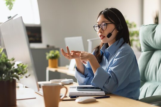 Focused Asian Female Customer Service Representative Wearing Headset, Communicating With Clients While Sitting At Modern Home Office Desk, Engaged In Work With Computer. Business Concept.