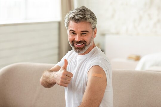 Smiling Gray Haired Man Giving Thumbs Up After Receiving Vaccine At Home, Feeling Happy And Safe. Displaying Arm With Bandage In Modern Living Room, Promoting Health Awareness.