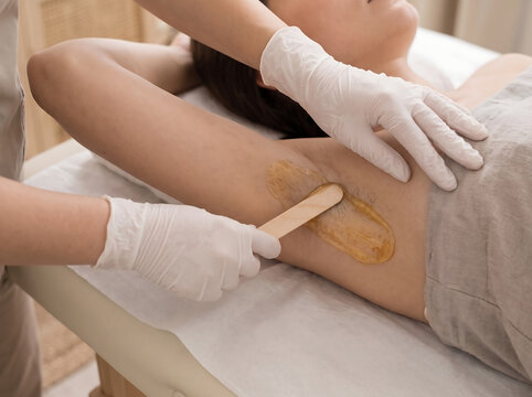 Beautician applying warm wax with wooden spatula on client armpit for underarm hair removal in spa