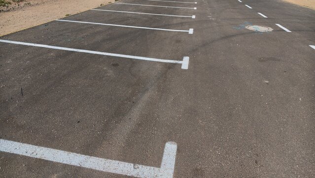 Empty asphalt parking lot with fresh white painted parking space lines, long tire marks on the dark gray surface, a round manhole cover and a sandy dirt edge along the side, and dashed lines far ahead