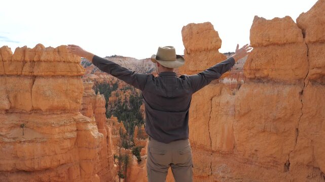Back view male hiker raising his arms in triumph while standing a top orange sandstone hoodoos and enjoying scenic canyon vista in bryce canyon national park