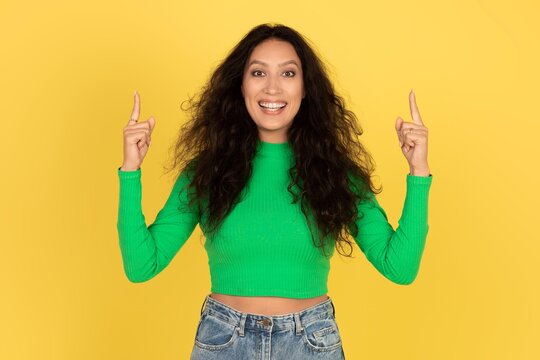 Smiling Young Woman With Curly Hair Gesturing With Both Hands Pointing Up At A Vibrant Yellow Background, Celebrating An Achievement, Capturing A Joyful Moment In A Studio Setting. Fun Concept.