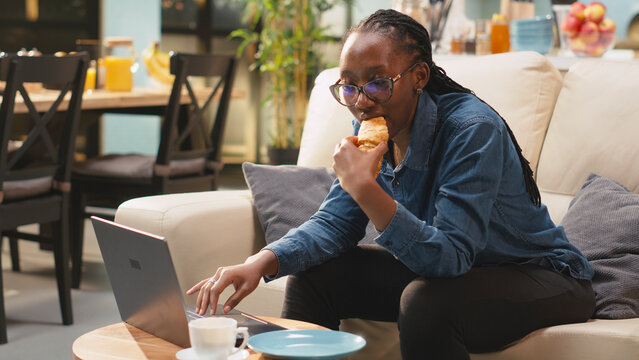 Croissant, laptop and snacking as woman eats pastry while working on computer from sofa at home. Remote work, multitasking and convenience shape casual mealtime and screen use in living room.