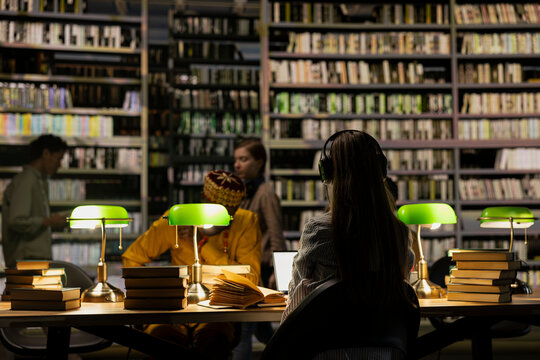 Headphones, library study and student as woman seen from behind works at table beneath green lamps with peers nearby. Focus, independent learning and concentration guide quiet academic preparation.