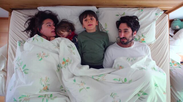 Top view of a Caucasian family of four, mother, father, son and toddler, lying side by side under a white duvet in bed, calm and cozy morning together.