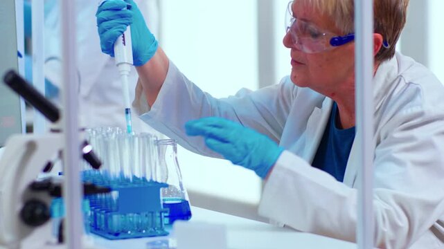 Micropipette, scientist and liquid analysis as an older woman measures solution into glassware beside test tubes and a microscope, for pharmaceutical research, precise dosing and sample testing.