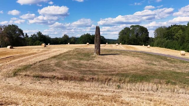 An impressive monolith, around 4000 years old, rises on a dry meadow. In the background, there is a harvested grain field with typical round bales of straw in front of a dense row of trees. The blue 