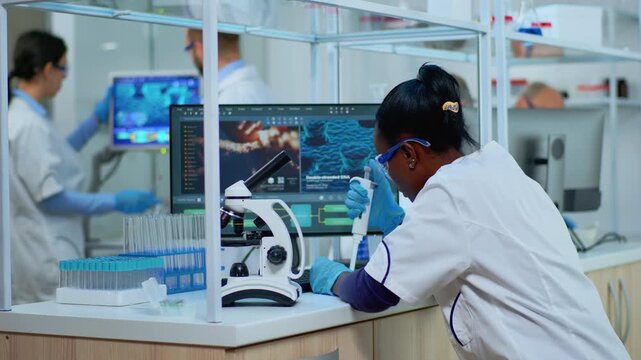 Pipette, scientist and microscope research as a woman scientist uses an instrument near the microscope while colleagues work behind her; precise sample handling, virology, biotechnology and testing.