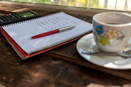 Close-up of budget notebook, red pen, and coffee cup on wooden table near window