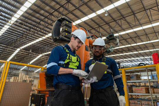 An industrial robot undergoes automation inspection while two coworkers review a laptop beside its orange arm inside a fenced cell.