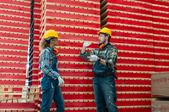 Wooden pallet logistics in the warehouse are discussed by two coworkers holding a tablet beside stacked red lumber.