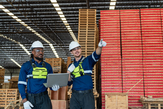 Workers checking lumber inventory in a warehouse use a laptop as one of the two men points above wooden pallets.