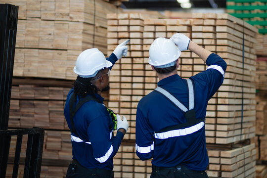 Workers checking lumber inventory in a warehouse stand beside each other as one points toward tall stacks of timber.