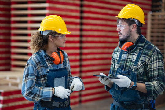 Female carpenter: woman discusses carpentry with her workshop colleague while holding a tablet in front of stacked lumber.
