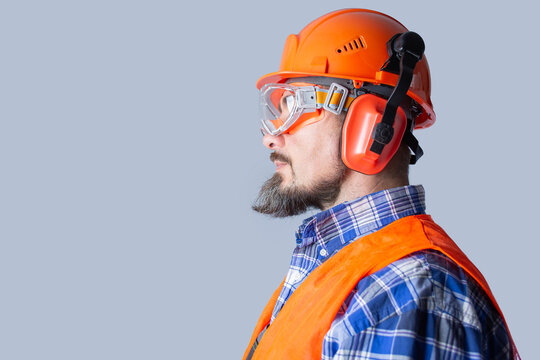 Worker in hard hat, safety goggles, earmuffs and orange vest on a gray background. Personal protective equipment and workplace safety for industrial training and recruitment banners with copy space.