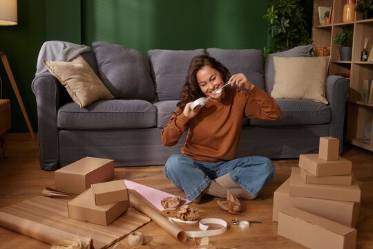 Smiling young woman sits on living room floor preparing to wrap cardboard gift boxes.