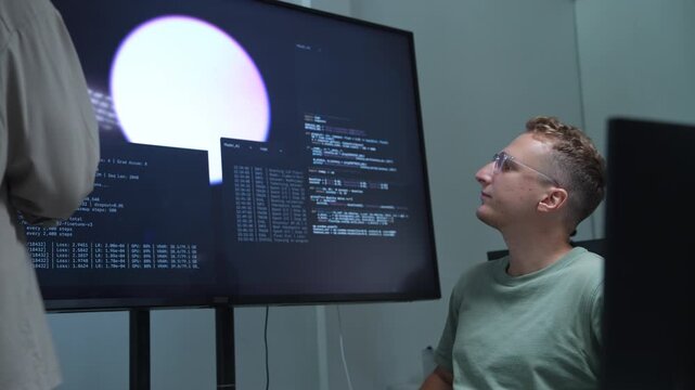Two AI engineers are collaborating in a modern workspace, discussing software development and digital data on a large computer screen for an advanced technology project.