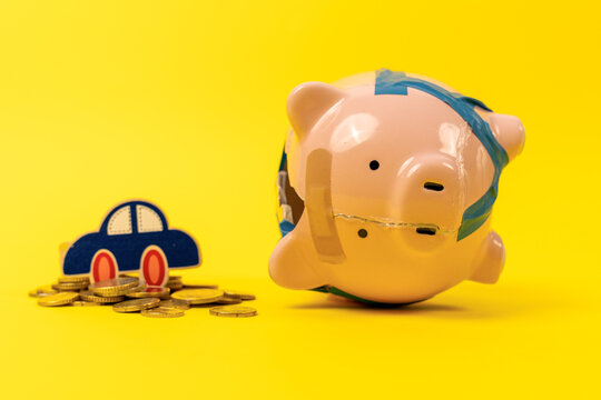 A pink piggy bank patched with sticky tape and a sticking plaster stands beside a blue paper car on a pile of golden coins against yellow, a concept of vehicle running expenses.