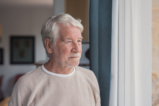 Elderly man with white hair and a beard, gazing thoughtfully through a bright window in his home, reflecting on aging, memory, retirement, and the quiet passage of time