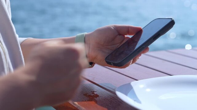 A woman in a light shirt and green wristband holds a smartphone and swipes the screen at a wooden table beside an empty white plate, with blue sea behind at an outdoor cafe.