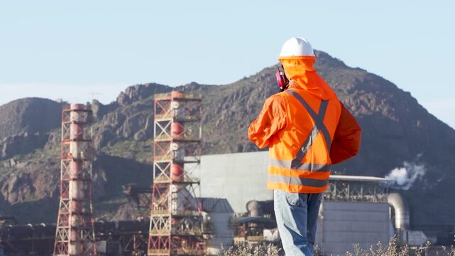 Industrial engineer wearing protective gear talking on a walkie talkie near an industrial plant