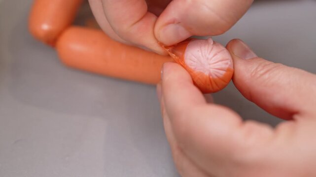A person pinches the end of an orange sausage and pulls away its transparent casing, exposing the pink meat underneath. More sausages rest nearby on a gray countertop, a concept of food preparation.