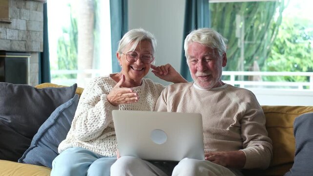 Senior couple sitting together on living room sofa, waving and smiling while making an online video call using portable computer device to connect with distant family