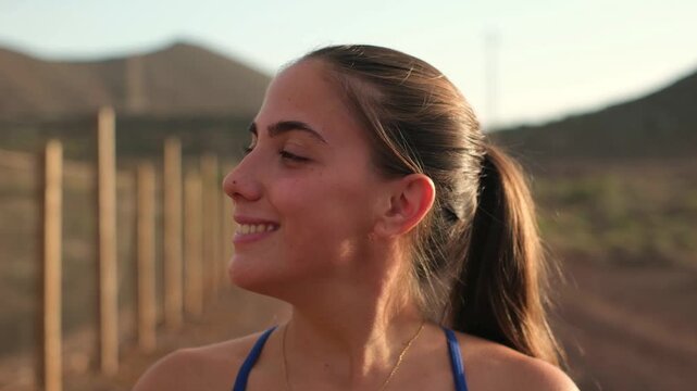 Attractive young female runner smiling and looking at the camera during golden hour, resting on a rural path by a wooden fence with hills in background