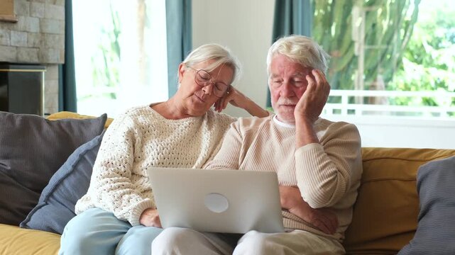 Stressed senior couple sitting on a cozy sofa at home, looking worried while checking online bills, banking problems, or bad financial news together on a modern laptop computer