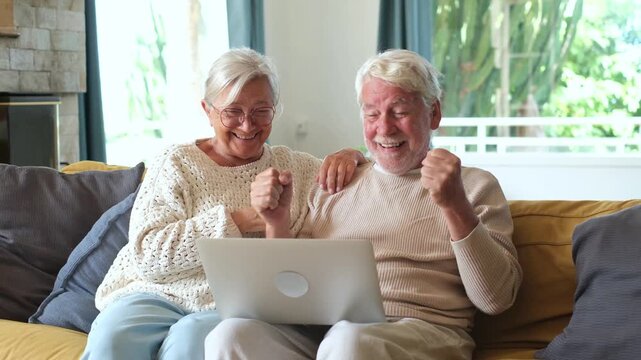 Joyful senior couple sitting on a cozy couch, celebrating good news on their laptop with raised fists and affectionate embraces, expressing happiness together in their living room