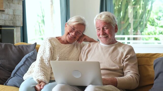 Smiling senior couple sitting together on a comfortable couch in living room, browsing internet and watching screen on modern laptop computer, enjoying leisure time