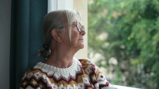Thoughtful senior woman wearing glasses and warm knitted sweater standing by window at home, looking outside into garden with pensive nostalgic expression during quiet afternoon